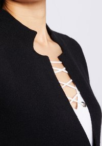 Czarny kardigan z wysokim kołnierzem i widocznym białym, krzyżowym sznurowaniem. Miękka faktura, minimalistyczny design i dopasowany krój.