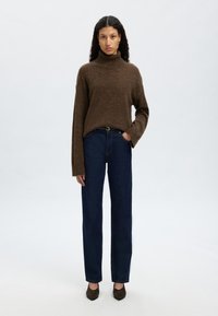 Brūns adījumu augstais kakls džemperis, brīvs piegriezums, apvienots ar tumši ziliem taisnām kājām džinsiem. Modelis valkā brūnas augstpapēžu kurpes. Vienkārša fona.