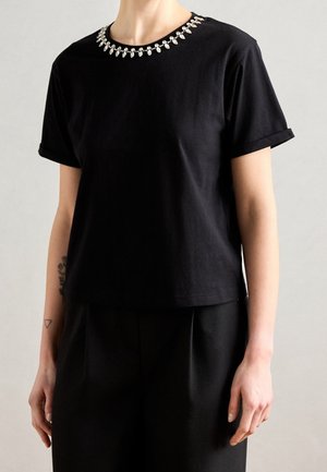 Persoon draagt een zwart shirt met korte mouwen met opgerolde manchetten en een ketting van heldere druppelvormige kralen rond de halslijn.
