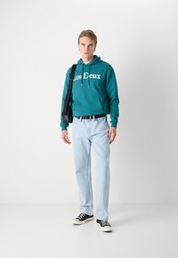 Teal hoodie with “Les Deux” text, kangaroo pocket, paired with light blue jeans and black canvas sneakers. Accessory: black shoulder bag.