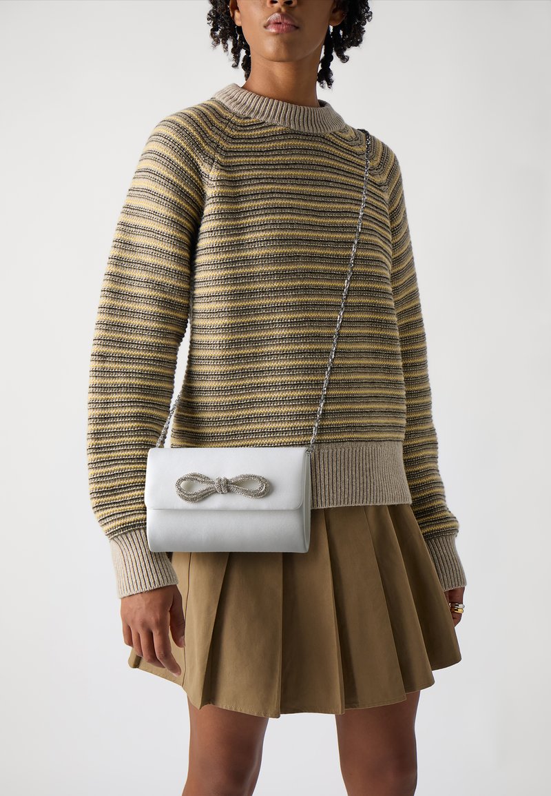 Geantă albă din piele, tip crossbody, cu un accent în formă de fundiță argintie, formă rectangulară și curea din lanț, purtată cu un pulover cu dungi și o fustă plisată.