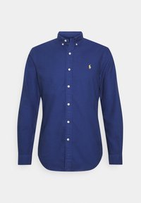 Pitkähihainen laivastonsininen napitettava paita, jossa on napitettava kaulus ja vasemmassa rinnassa pieni keltainen kirjailtu poloilijan logo.