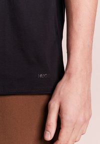 Camisa negra de manga corta con una suave textura de algodón. Presenta el logo "HUGO" en una impresión sutil cerca del dobladillo. Tela marrón visible debajo.