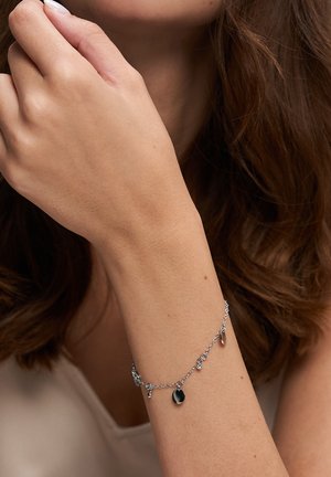 Woman's arm and hand near face, wearing a thin silver charm bracelet with small circular and crystal pendants.