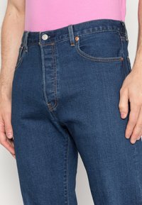 Jeans de ganga azul escuro com costura visível, design clássico de cinco bolsos e fecho frontal com botão. Feitos de um tecido robusto e texturizado.