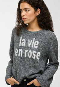 Frau mit lockigem, dunklem Haar, die einen grauen Strickpullover mit weißem Text "la vie en rose" trägt, die Hände in den schwarzen Hosen Taschen, schaut zur Seite.