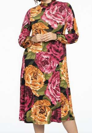 Femme portant une robe à manches longues, longueur genou, avec un grand motif floral orange et rose sur fond noir. Mains jointes à la taille.