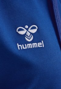 Blauwe textiel met een geborduurd wit logo met symmetrische vormen en de tekst "hummel", met een gladde textuur.
