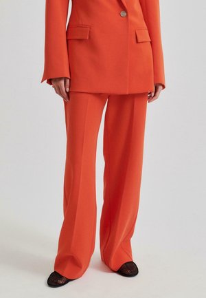Kobieta w pomarańczowym, dopasowanym garniturze z szerokimi spodniami i dwurzędową marynarką, w połączeniu z czarnymi, siateczkowymi butami na szpic.