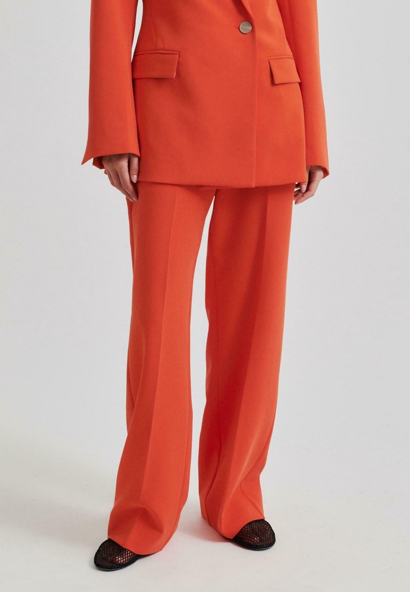 Femme portant un costume sur mesure orange avec un pantalon à jambes larges et un blazer à double bouton, assorti à des chaussures noires en maille à bout pointu.