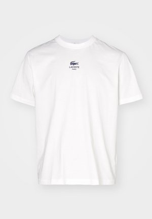 Hvid T-shirt med korte ærmer, rund hals og lille sort Lacoste-logo samt teksten "LACOSTE PARIS" centreret på brystet.