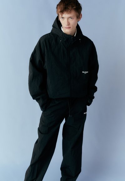 Black jacket and pants set with a textured finish, featuring a zippered front, an adjustable hood, and subtle white branding on both pieces.