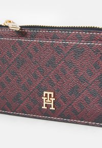 Red and black patterned wallet in textured material, featuring a gold logo and a zip closure with gold hardware. White stitching accents.