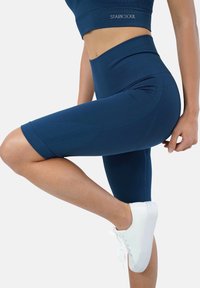 Leggings high-waisted blu navy con un design testurizzato, taglio a metà gamba e un sottile logo sulla vita. Abbinati a scarpe da ginnastica bianche.