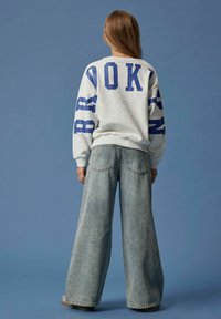 Person som står med ryggen mot kameran, iklädd en grå sweatshirt med blå texten "BROOKLYN" och vida ljusblå jeans mot en blå bakgrund.