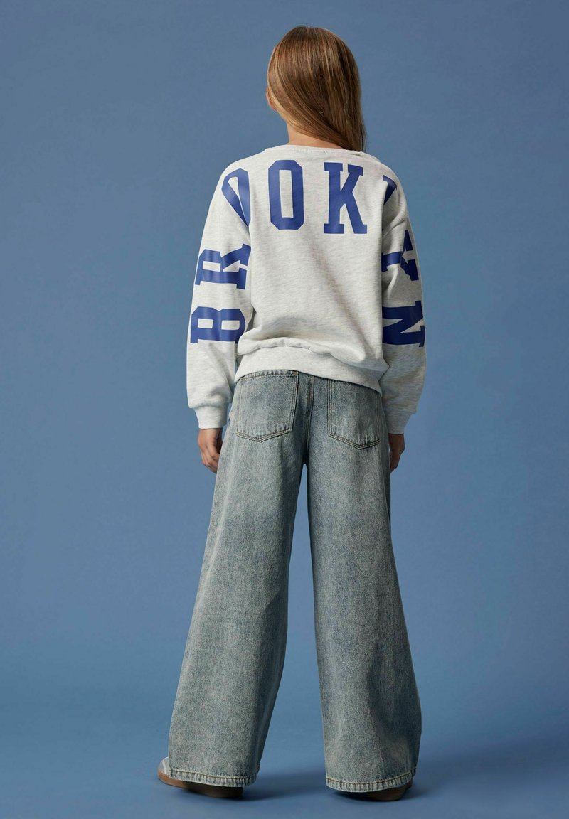 Person som står med ryggen mot kameran, iklädd en grå sweatshirt med blå texten "BROOKLYN" och vida ljusblå jeans mot en blå bakgrund.
