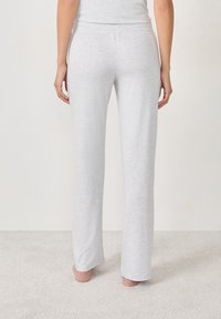 Persoon staat blootsvoets op tapijt, draagt lichtgrijze aangesloten loungebroeken, gezien van achteren tegen een neutrale muur.