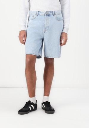 Light blue denim shorts with a straight cut, five pockets, and frayed hem. Paired with black sneakers featuring white stripes.