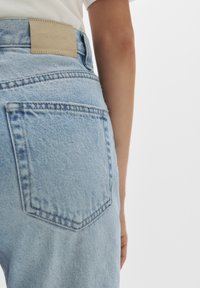 Világoskék farmernadrág középmagas derékkal, bézs bőr logópemzzel, két hátsó zsebbel és enyhén laza szabással.