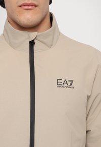 Beigeová bunda s vysokým golierom, čiernym zipsom a logom EA7 Emporio Armani na hrudi. Hladká textúra a ľahký materiál.