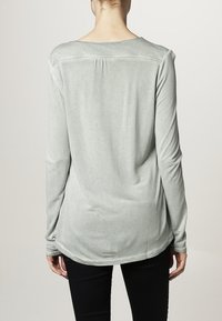 Maglia a maniche lunghe di colore grigio chiaro con orlo arrotondato, vestibilità leggermente ampia e texture morbida; presenta un dettaglio di cucitura lungo il colletto posteriore.