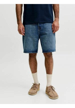 Homme portant un short en jean bleu mi-long, des chaussures en daim beige à lacets, des chaussettes couleur crème et une chemise bleu marine foncé, debout les bras détendus.