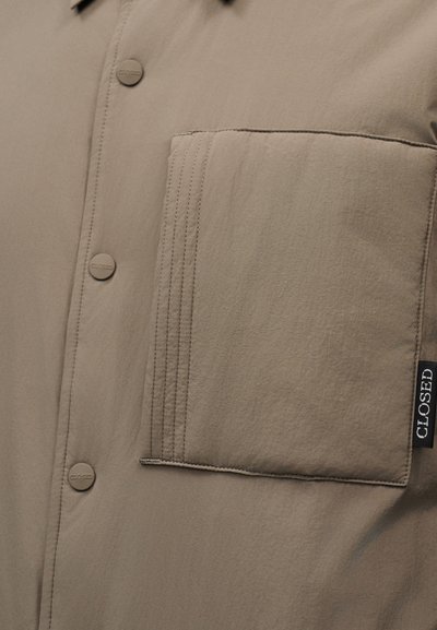 Κοντινό πλάνο από το εμπρός μέρος ενός μπεζ πουκαμίσου που δείχνει μια μεγάλη ορθογώνια τσέπη στο στήθος με ραμμένες λεπτομέρειες και κουμπιά με το λογότυπο κατά μήκος της πατιλέτας.