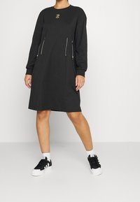 Svart långärmad klänning med rund halsringning, sidogömmor och guldblad broderi vid bröstet. Matchad med svart och vitt sneakers.