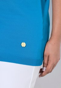 Blaues gestricktes Oberteil mit einem gerippten Saum und einem goldenen Logo-Akzent an der Seite, kombiniert mit einem weißen Stoff, was den Textur- und Farbkontrast hervorhebt.