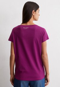 Achteraanzicht van een paarse, kortslachtige t-shirt gemaakt van zachte katoen; voorzien van een ronde halslijn en een logo gedrukt nabij de schouder.