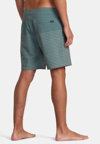 Bañadores de hombre en un color teal apagado con rayas horizontales blancas, hechos de material ligero. Cuenta con un bolsillo trasero y una cintura elástica.
