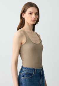 Vrouw met lang bruin haar, gekleed in een beige geribbelde tanktop en blauwe jeans, staand tegen een effen lichtgrijze achtergrond.