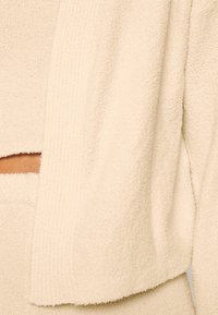 Tessuto di un bianco sporco con una struttura texturizzata, caratterizzato da un design morbido e dettagli a costo. Il tessuto appare morbido, con una sensazione accogliente e soffice. Dettaglio a forma di taglio visibile.