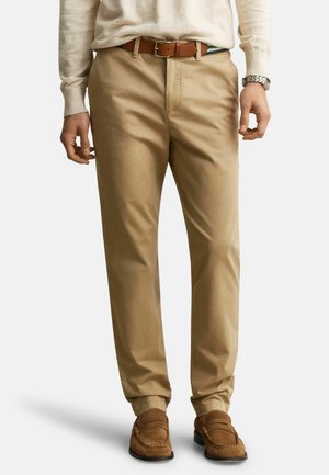 Homme portant un pantalon chino beige, une ceinture en cuir marron, un pull blanc, des mocassins en daim marron et une montre-bracelet argentée, debout devant un fond uni.