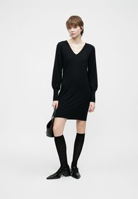 Čierne pletené šaty s V-výstrihom, dlhými puff rukávmi a priliehavým strihom, doplnené nadkolenkovými ponožkami a čiernymi topánkami.