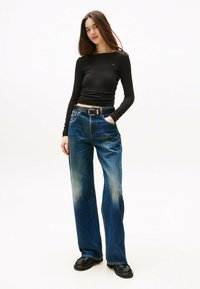 Schwarzes Langarmoberteil mit Rundhalsausschnitt, kombiniert mit blauen, weiten Denim-Jeans mit ausgewaschenen Stellen und einem schwarzen Gürtel. Schwarze Schuhe.