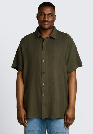 Mann trägt ein dunkelgrünes kurzärmeliges Hemd mit Knöpfen und hellblaue Jeans, steht vor einem schlichten hellen Hintergrund.