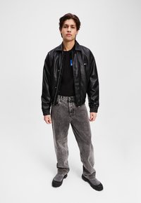 Svart skinnbomberjakke med elastiske mansjetter, kombinert med falmede grå vide jeans og svarte og grå støvler, satt mot en ensfarget bakgrunn.