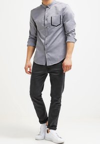 Camisa de botones gris claro con un bolsillo delineado en negro, combinada con pantalones gris oscuro y zapatillas blancas. Corte casual con mangas remangadas.