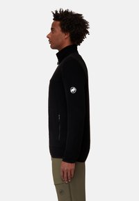 Giacca in pile nera con colletto alto, taschino con zip sul petto e logo ricamato sulla manica sinistra. Tessuto liscio, design aderente.