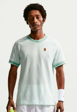 Mand iført lyse mintgrøn tennisshirt og håndledsbånd, der holder en gul tennisbold, stående mod ensfarvet baggrund.