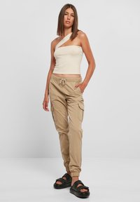 Beżowy krótki top na jedno ramię, zestawiony z jasnokawowymi spodniami cargo z bocznymi kieszeniami i elastycznymi ściągaczami. Czarne klapki.