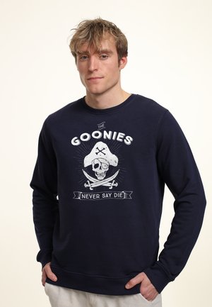Giovane uomo che indossa una felpa blu Navy con una grafica di teschio e spade incrociate e la scritta "The Goonies Never Say Die."