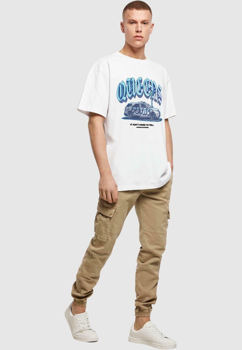 Hvit oversized t-skjorte med blå grafisk tekst "QUEENS" og illustrasjon, kombinert med beige cargo-bukser og hvite joggesko.