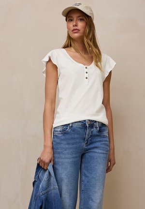 Jeune femme portant une casquette beige, un haut blanc à manches froufroutantes avec des boutons, un jean bleu, tenant une veste en denim sur fond beige.