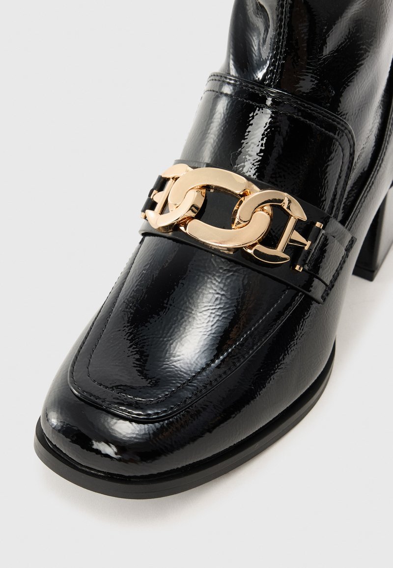 Mocasines de charol negro con un acento de cadena dorada en la parte superior, que presentan una punta redondeada y un tacón cuadrado para mayor estabilidad.