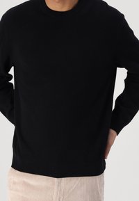 Mann in einem schlichten schwarzen Rippenpullover mit langen Ärmeln und beigen Cordhosen, die Hände auf dem unteren Rücken abgestützt.