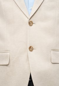 Μπεζ σακάκι με δύο καφέ κουμπιά πάνω από ένα ανοιχτό γαλάζιο πουκάμισο, με εμφανείς τις μπροστινές τσέπες και μέρος μιας σκούρας ζώνης και παντελονιού.