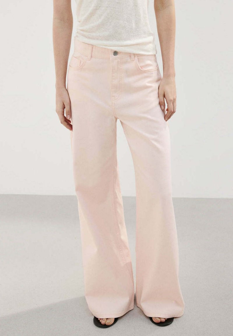 Pantalones anchos de pierna de color rosa claro, usados con una camiseta blanca metida y sandalias negras de punta abierta, mostrados en una persona de pie.