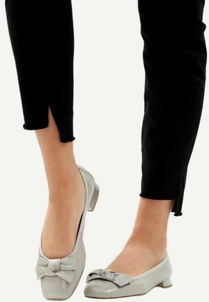 Pieds chaussés de chaussures gris clair à petits talons ornées de nœuds, associées à un pantalon noir avec de petites fentes à l'avant au niveau des chevilles.